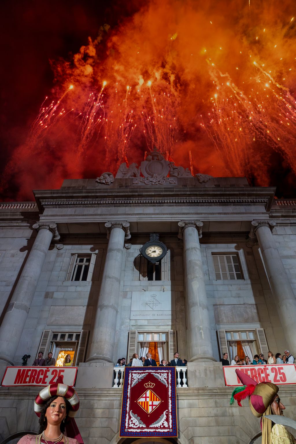 Focs artificials a la façana de l’Ajuntament, com a toc d’inici de la Mercè 2025, mentre els representants polítics i els mitjans de comunicació els miren des del balcó de l’Ajuntament